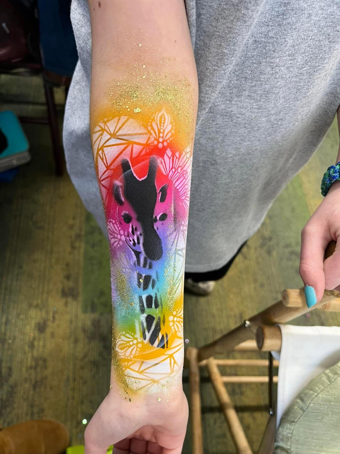 Rainbow airbrush giraffe design painted along a child's forearm at an outdoor event