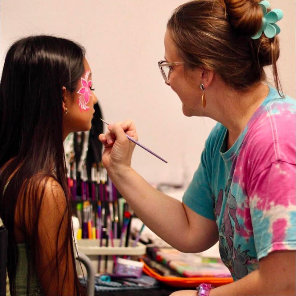 Pink hibiscus flower face paint being applied above a client's eye by face painter Tiah