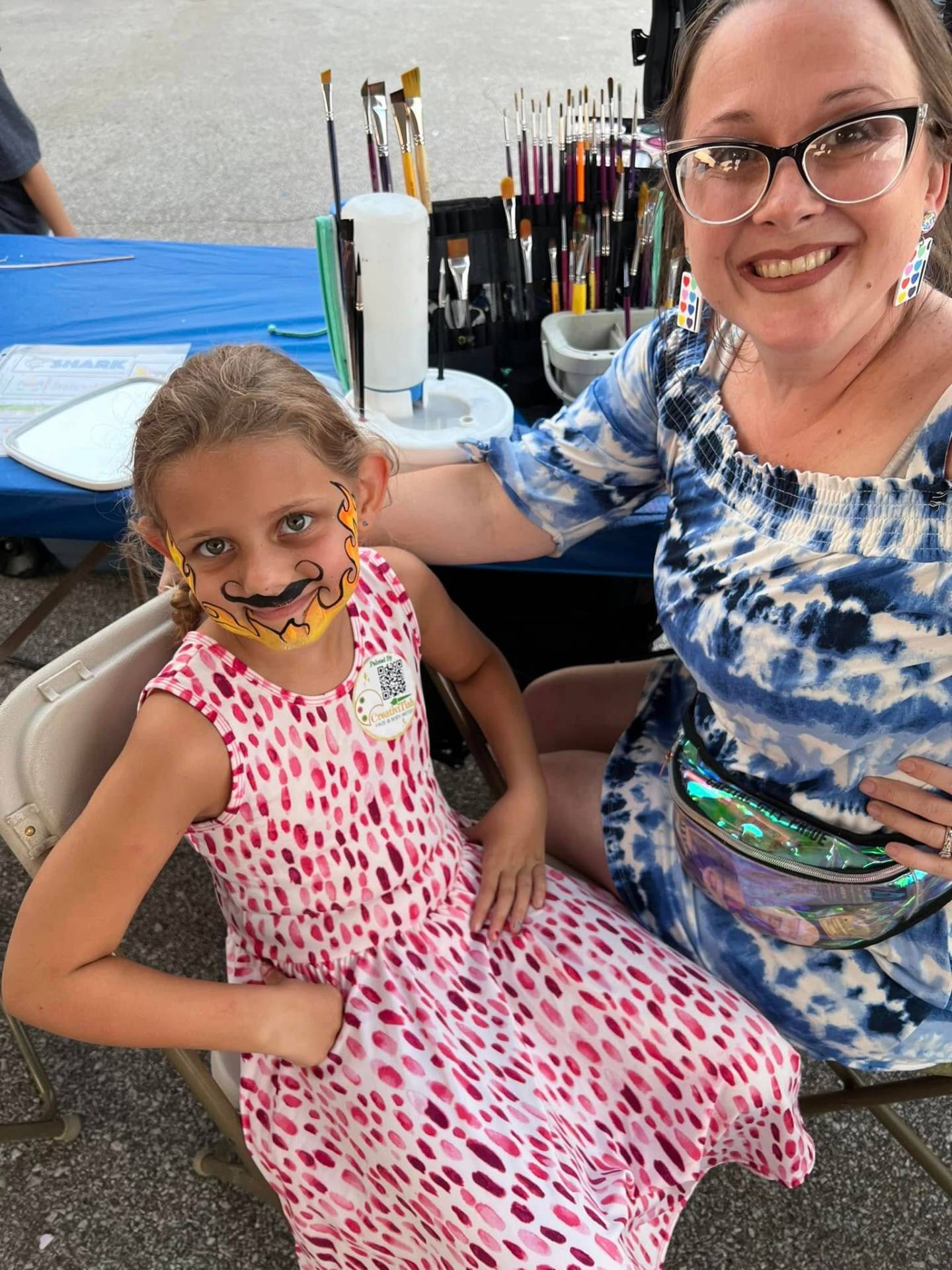Tiah, professional face and body artist, painting at an outdoor event with a happy child showing off her flame design